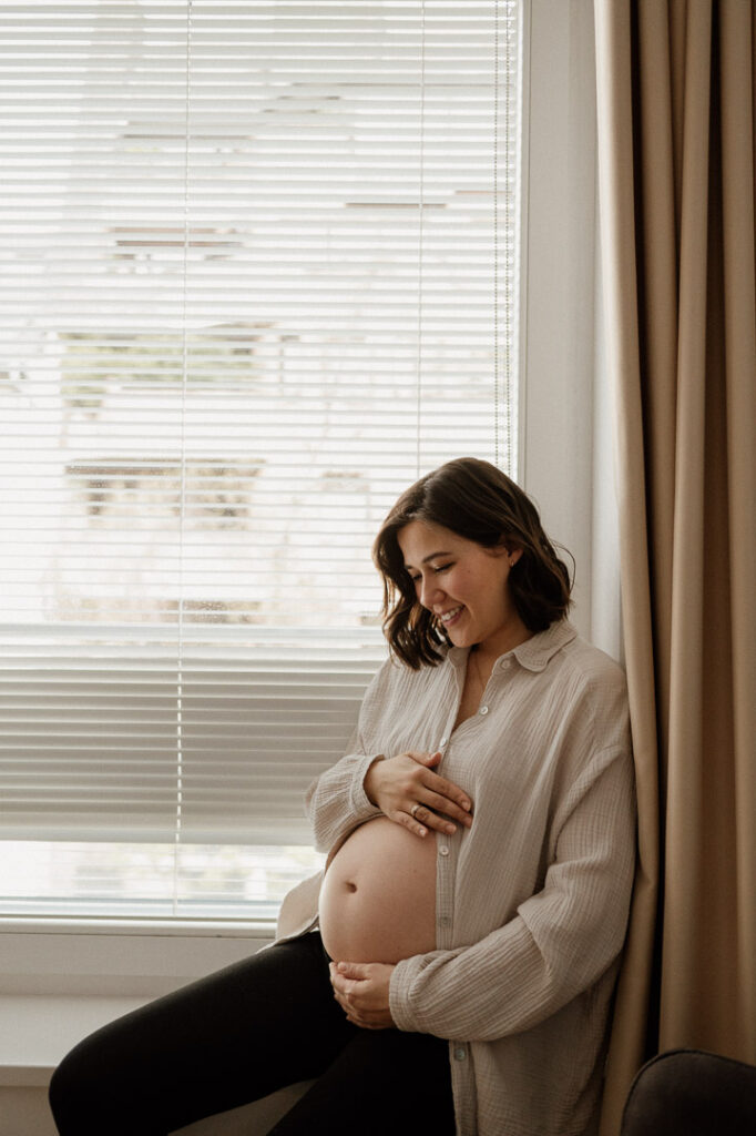 Schwangere sitzt bauchfrei auf Fensterbank